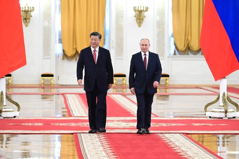 putin og xi.jpg To menn i dress kjem gåande mot kamera i ein stor sal. Flagg, tepper og gullpynt om omgjevnadene.